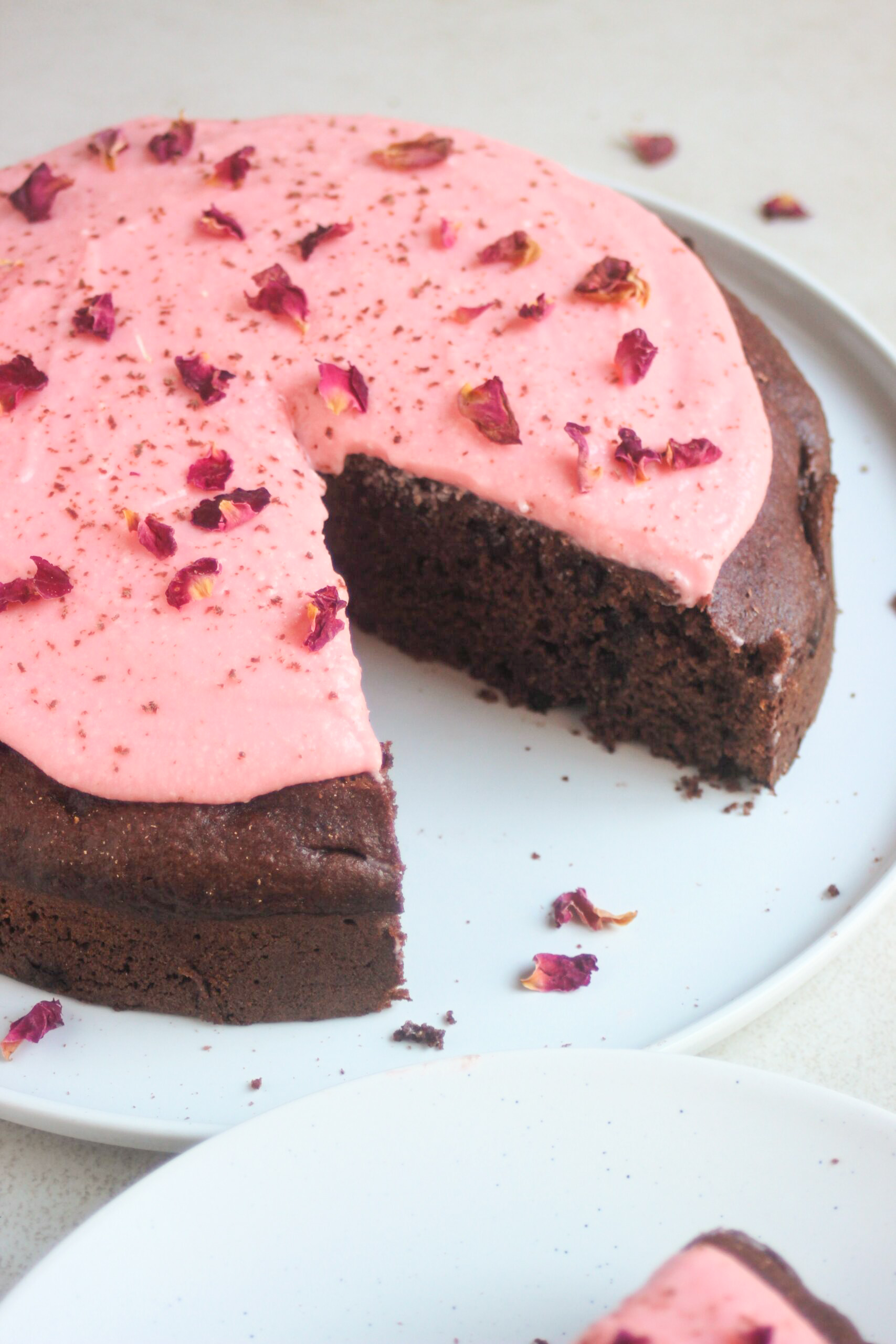 Chocolate beet cake with pink frosting and rose leaves, without a portion, on a white plate.