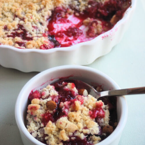 Small bowl with plum crumble and a spoon. Whole crumble behind on a baking dish.
