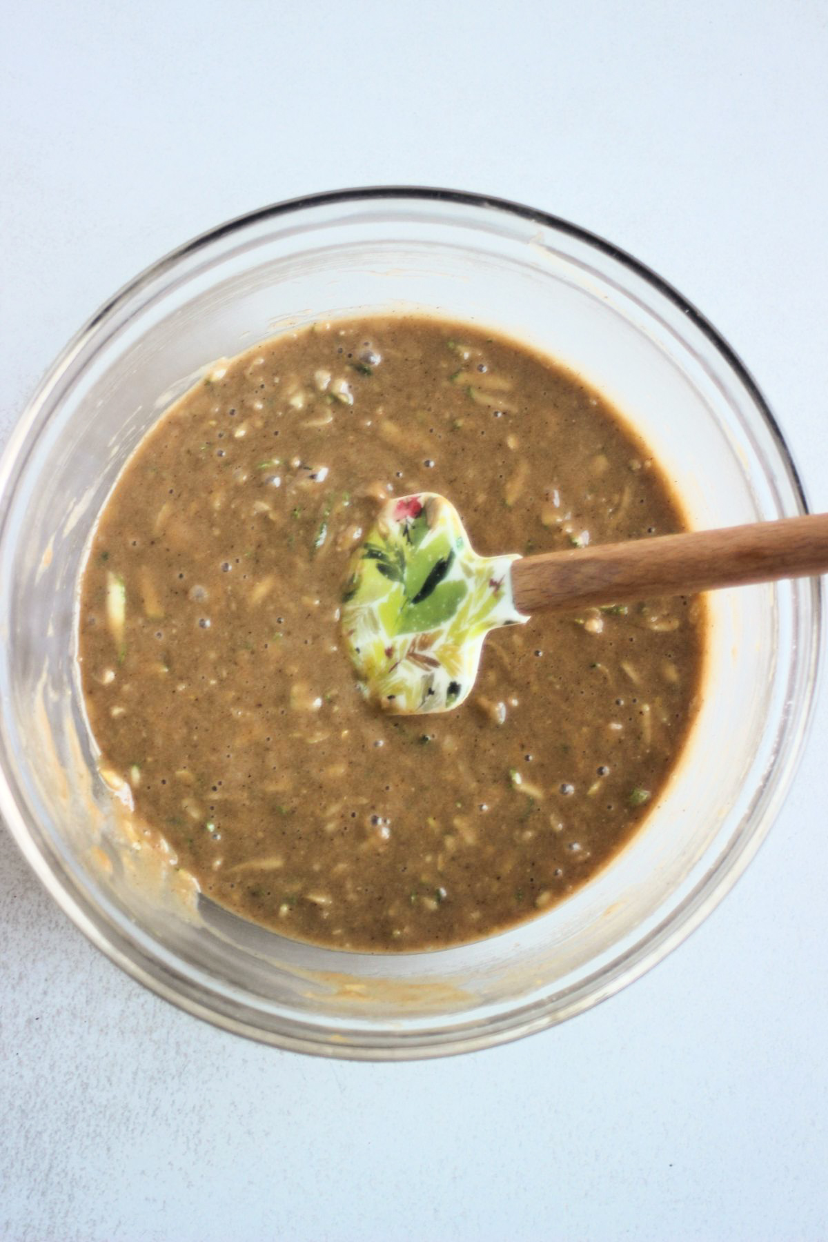 Glass bowl with a light brown mixture that includes grated zucchinis and a rubber spatula.