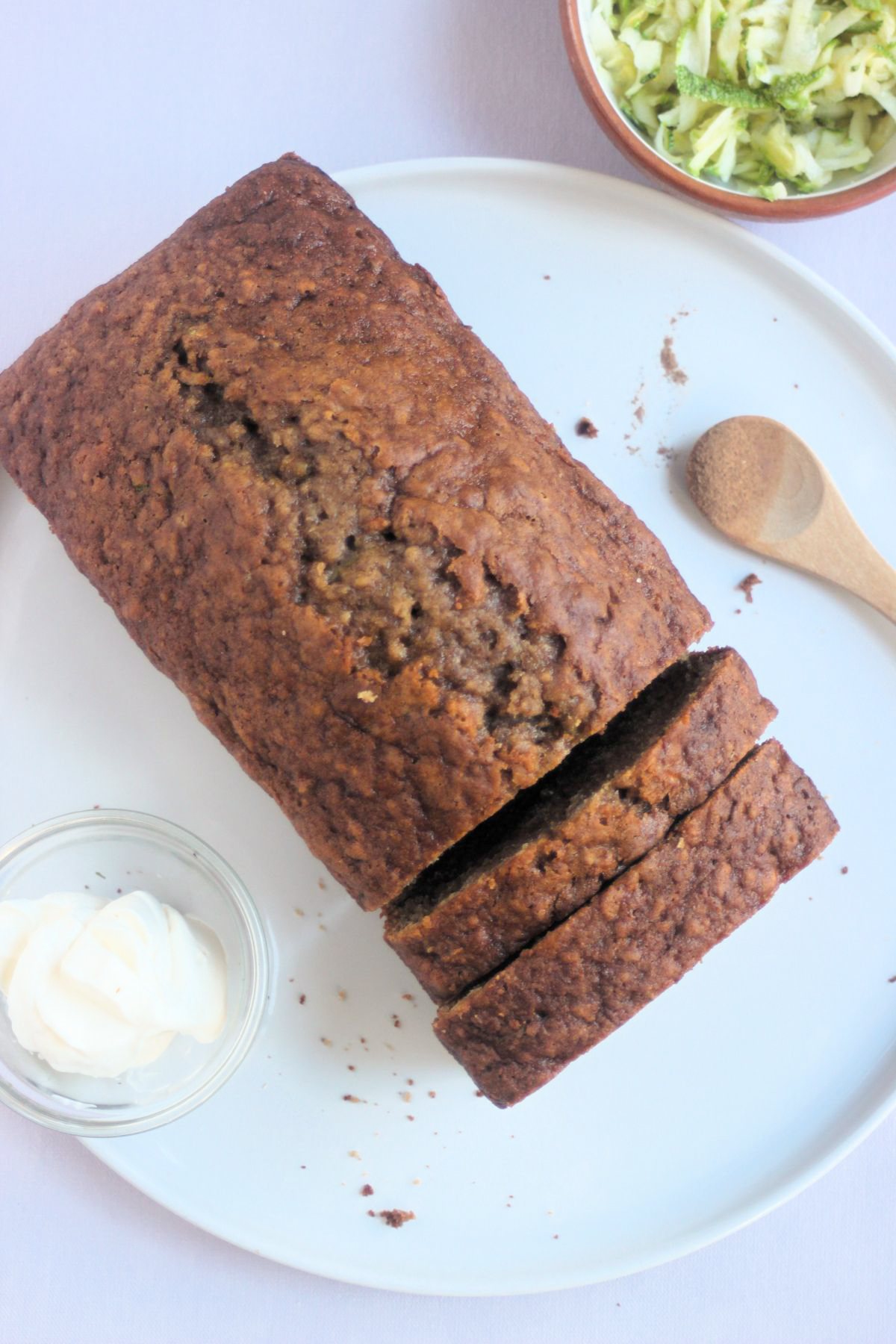 Top view of zucchini bread with two slices of it on a white plate. Tiny bowl with cream cheese and a wooden spoon.