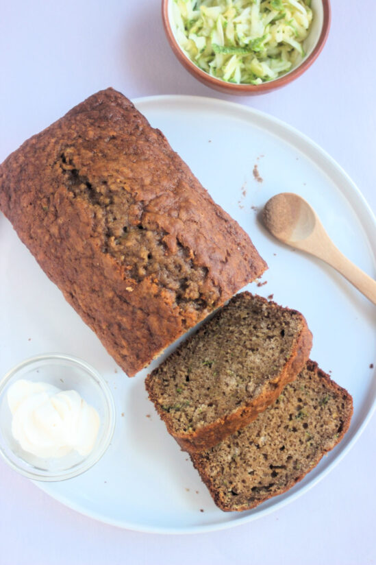Top view of zucchini bread and two slices of it on a white plate. Tiny bowl with cream cheese and a wooden spoon.