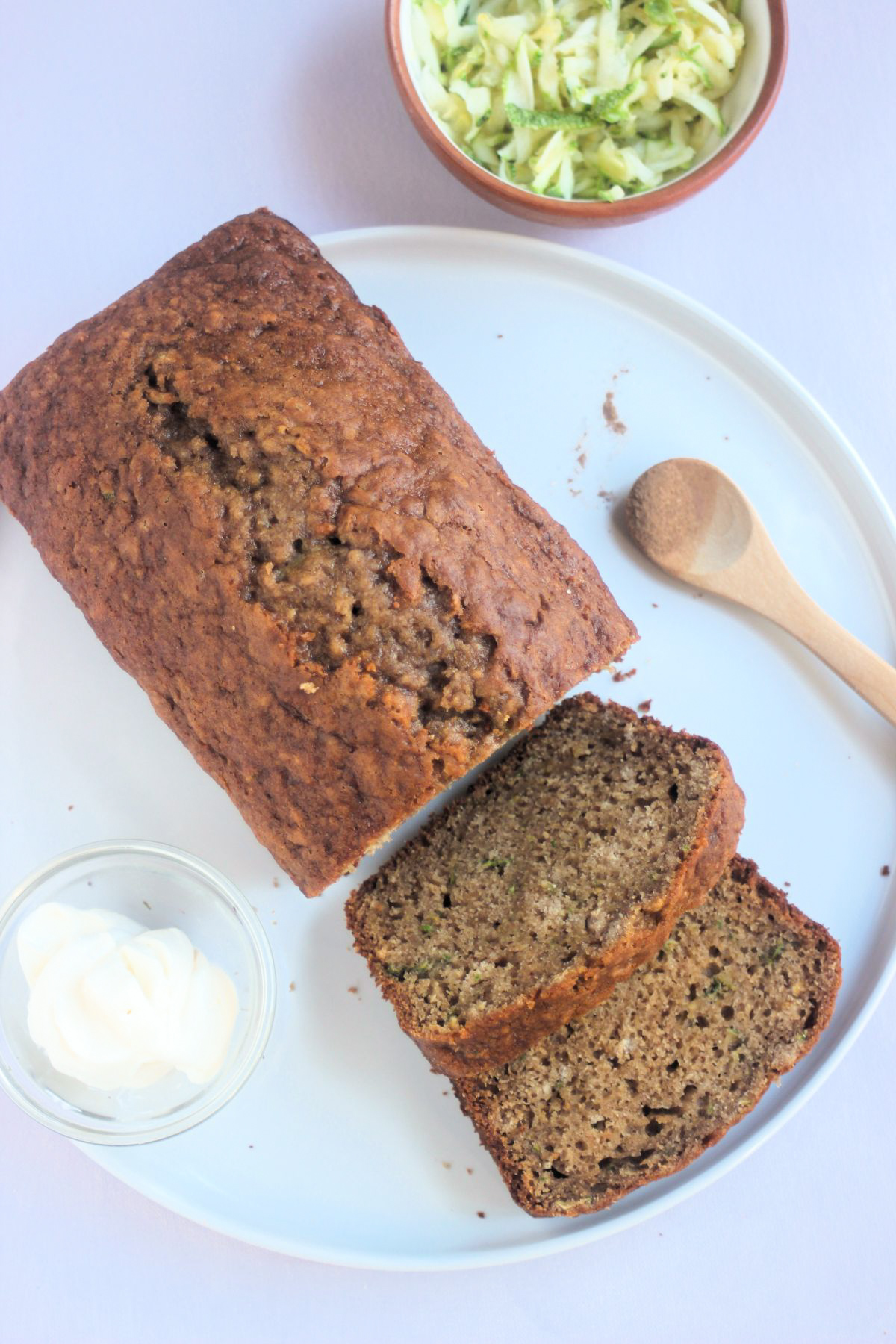 Top view of zucchini bread and two slices of it on a white plate. Tiny bowl with cream cheese and a wooden spoon.