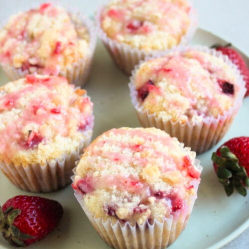 Strawberry streusel muffins with liners and strawberry icing on an aqua green plate. Fresh strawberries on the sides.