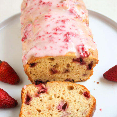 Strawberry bread with pink icing, and a portion of it on a white plate. Fresh strawberries on the sides.