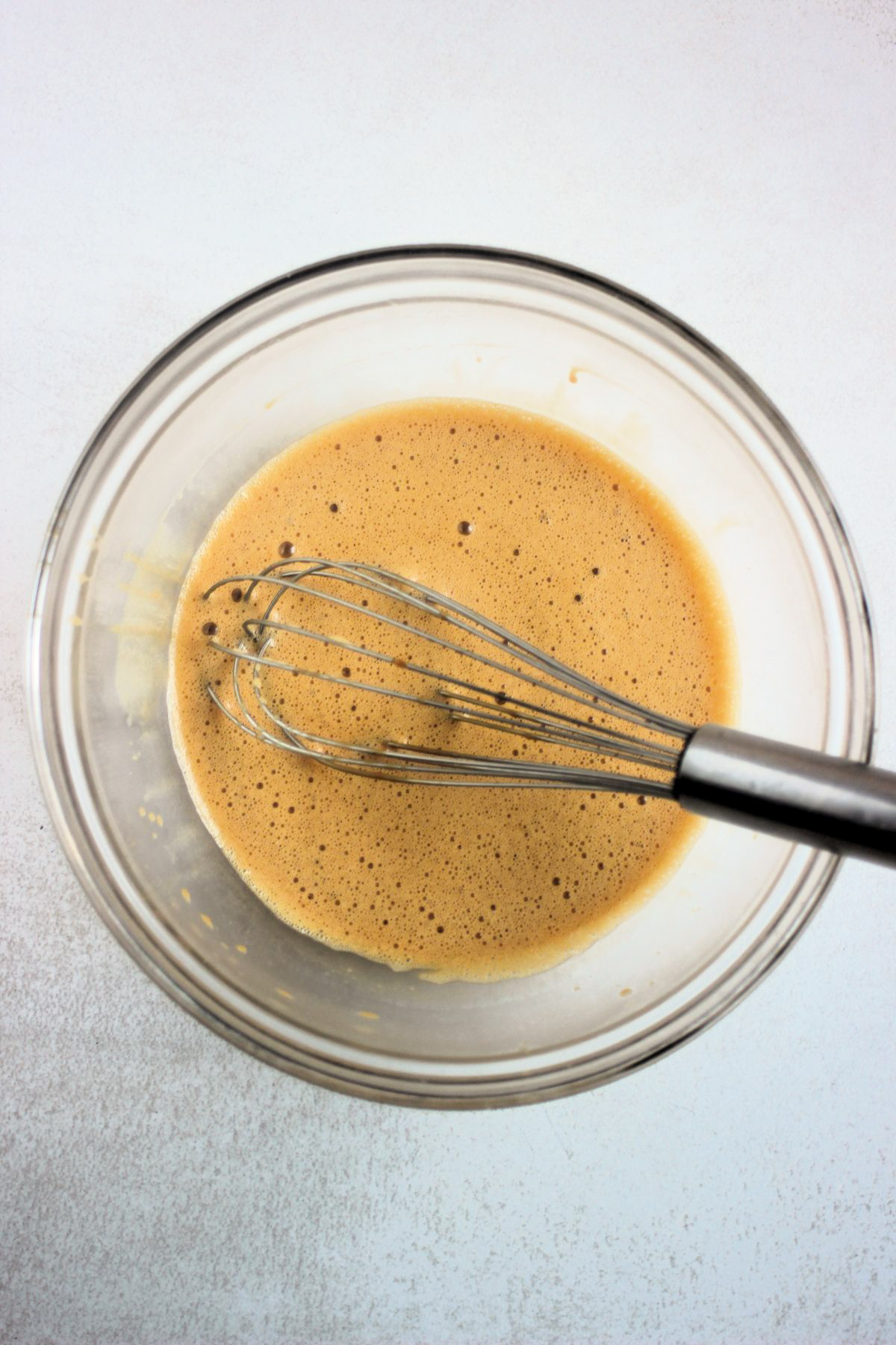 Glass bowl with a caramel-colored liquid and a hand whisk.