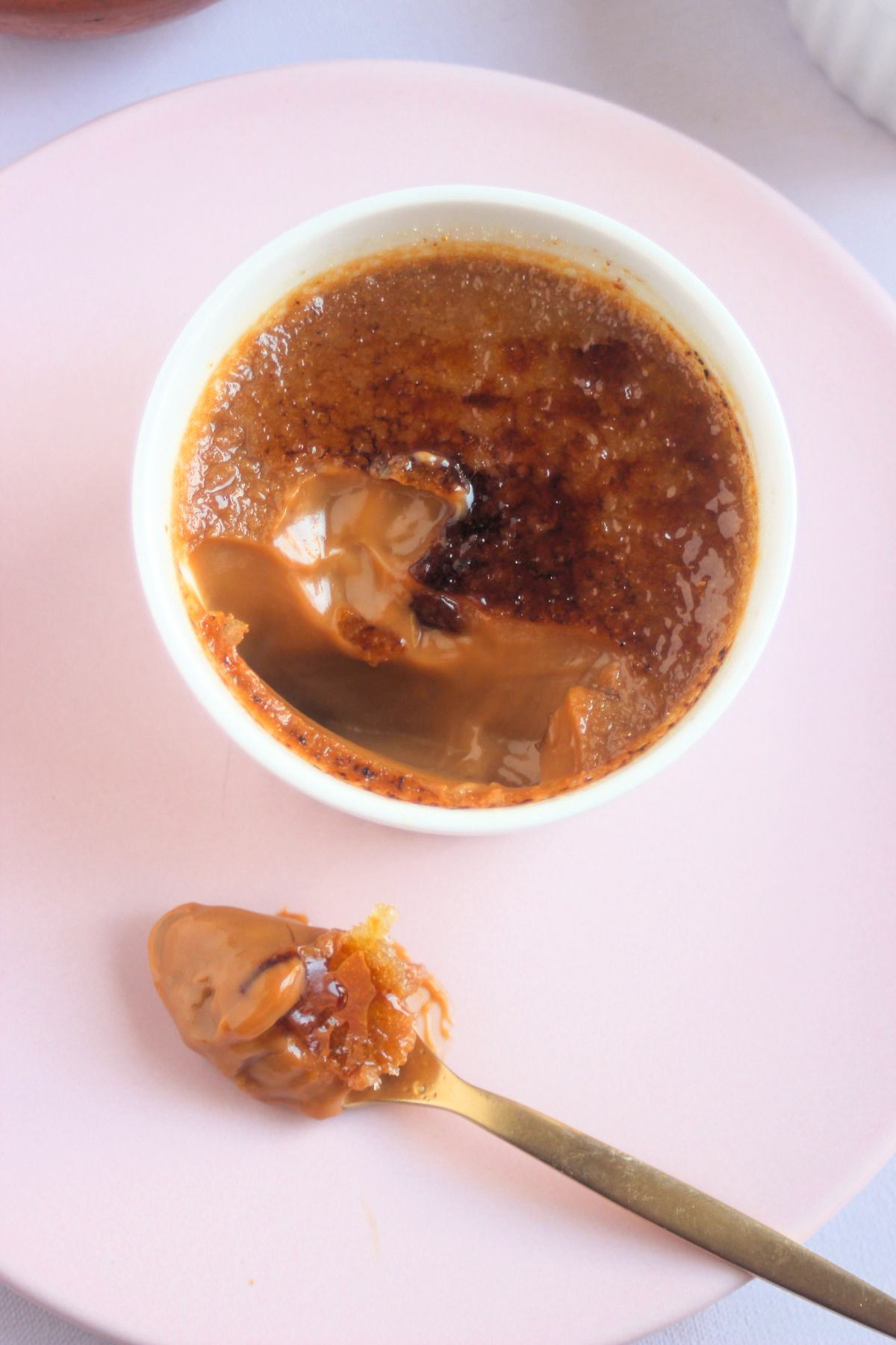 Top view of a partially eaten crème brûlée on a pink plate and a spoon with crème brûlée.