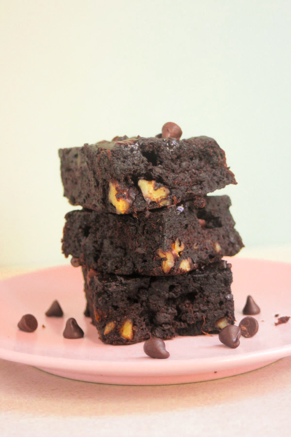 Front view of a stack of three sweet potato brownies on a pink plate. Chocolate chips are scattered on the plate.