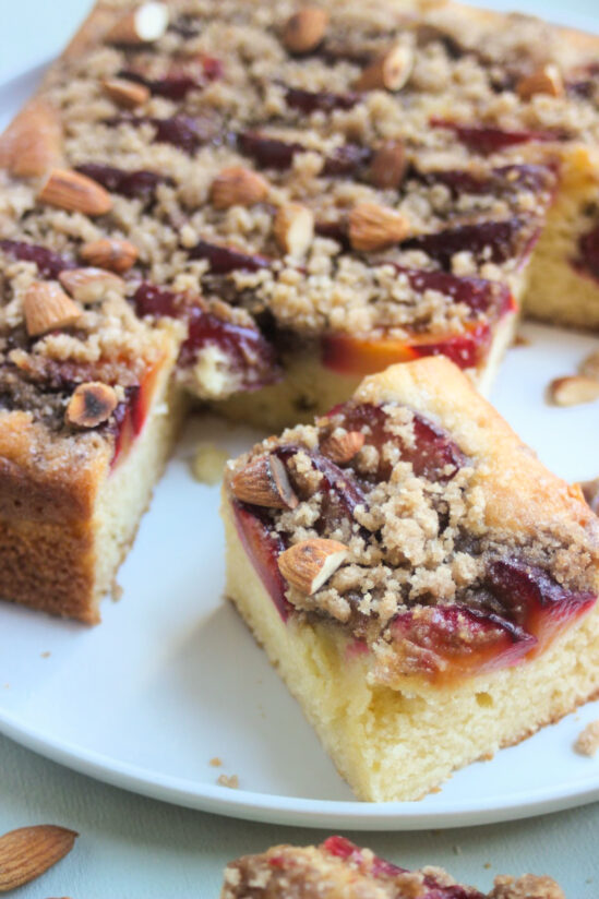 Portion of plum crumb cake on and the cake behind on a white plate.