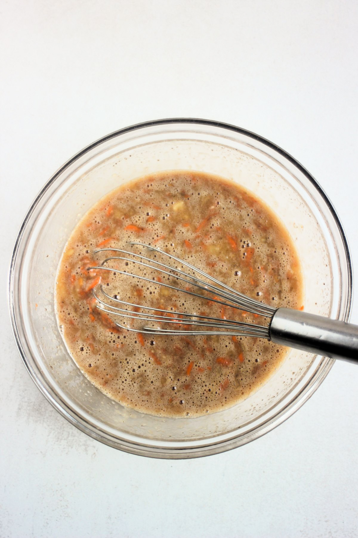Glass bowl with a brown liquid mixture that includes grated carrots and a hand whisk.