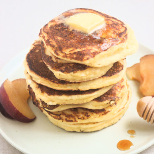 Stack of cottage cheese pancakes with a cube of butter on top and fresh plum slices on the sides on a white plate.