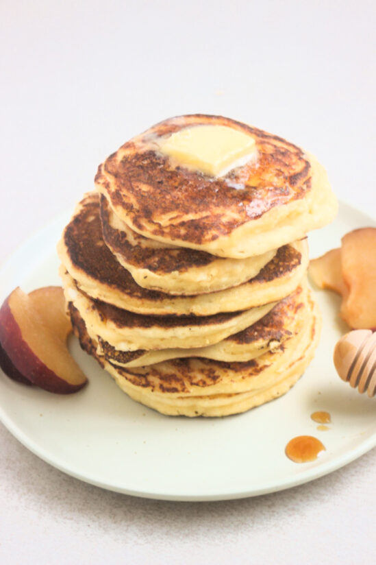 Stack of cottage cheese pancakes with a cube of butter on top and fresh plum slices on the sides on a white plate.