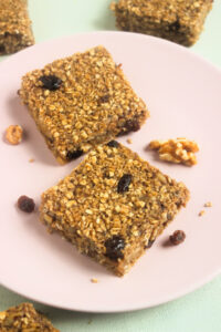 Two baked oatmeal bars on a pink plate. Raisins and walnuts are scattered around.