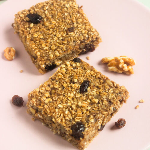 Two baked oatmeal bars on a pink plate. Raisins and walnuts are scattered around.