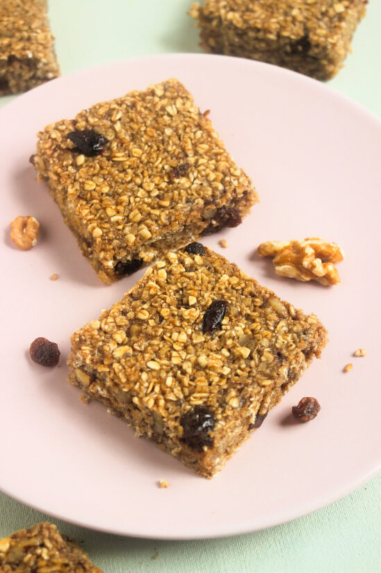 Two baked oatmeal bars on a pink plate. Raisins and walnuts are scattered around.