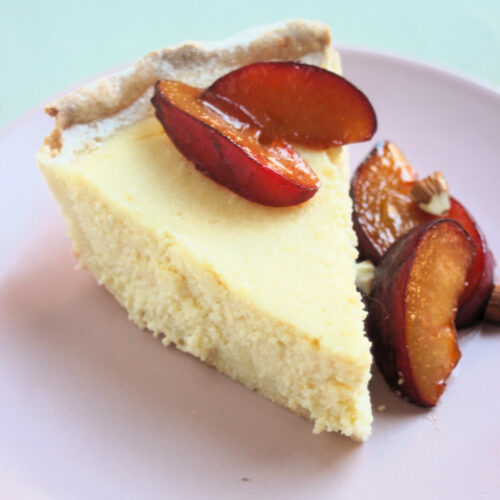 Portion of ricotta pie on a pink plate with plums.