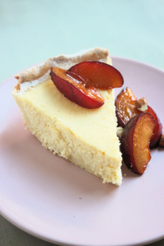 Portion of ricotta pie on a pink plate with plums.