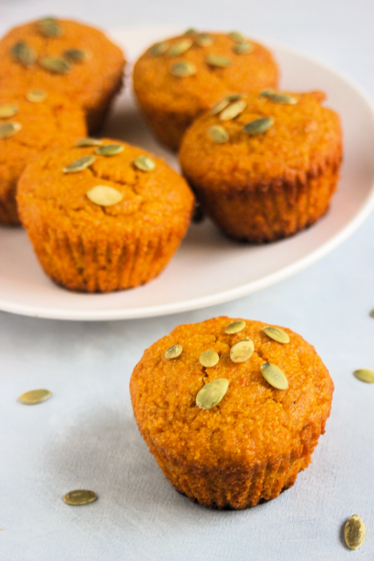 Pumpkin cornbread muffin with pumpkin seeds on a white surface. More muffins behind on a white plate.