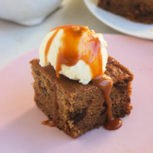 Portion of date cake topped with a scoop of ice cream and caramel sauce on a pink plate.
