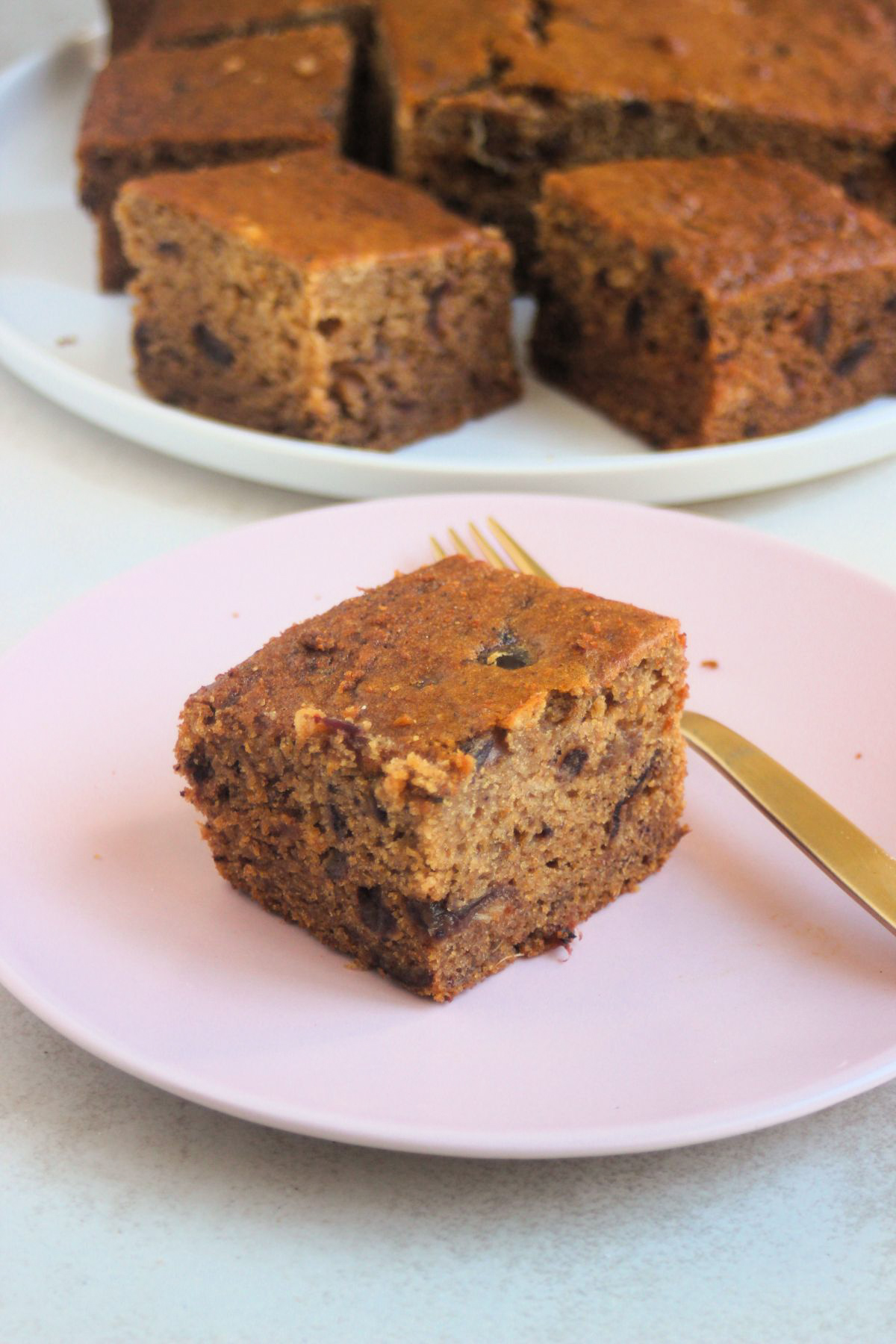Portion of date cake and a golden fork on a pink plate. More cakes are behind on a white plate.