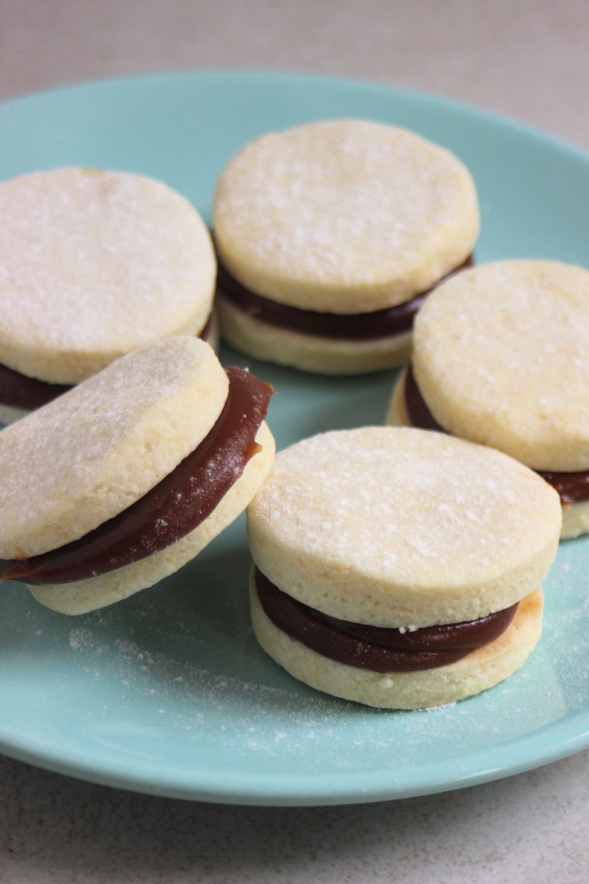 Almond alfajores on a green aqua plate. One alfajor is resting on another alfajor.