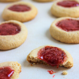 Almond thumbprint cookies on a white surface. One is broken, and the strawberry jam falls off.