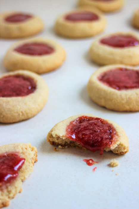 Almond thumbprint cookies on a white surface. One is broken, and the strawberry jam falls off.