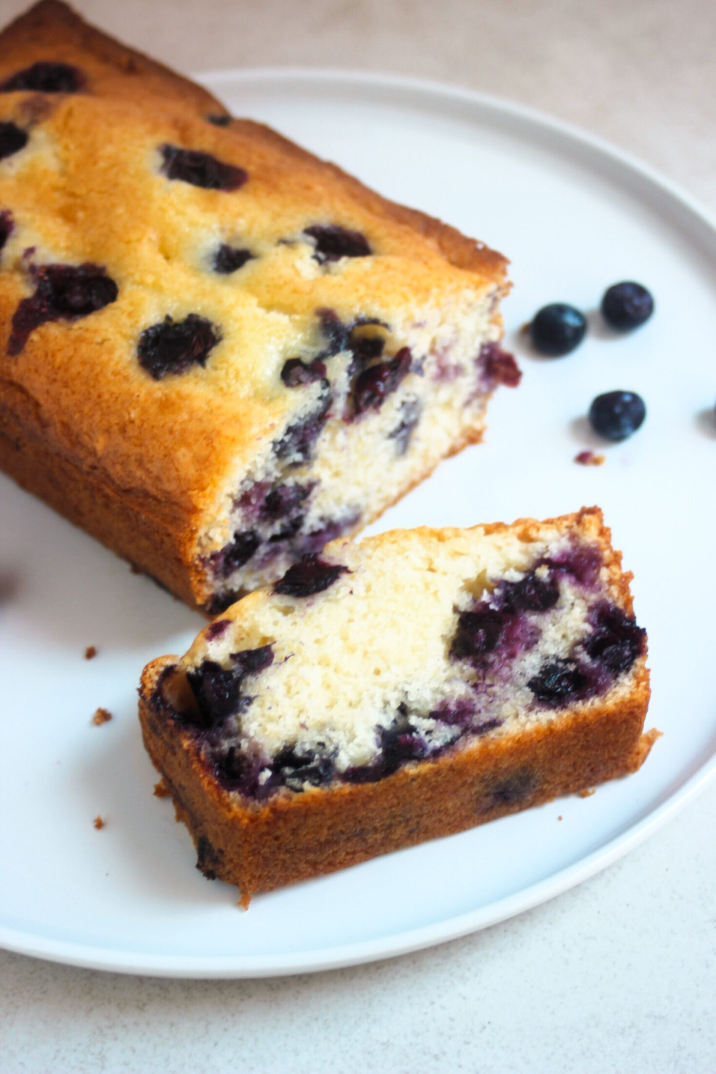 Blueberry loaf cake and a slice of it on a white plate. Fresh blueberries on the sides.