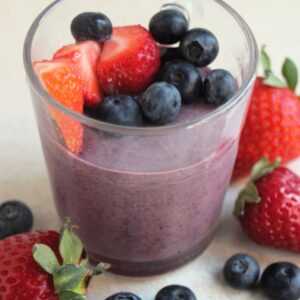 Glass cup with blueberry panna cotta, blueberries, and strawberries. More strawberries and blueberries are scattered around.