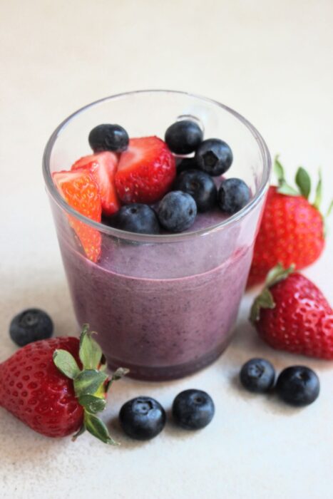 Glass cup with blueberry panna cotta, blueberries, and strawberries. More strawberries and blueberries are scattered around.