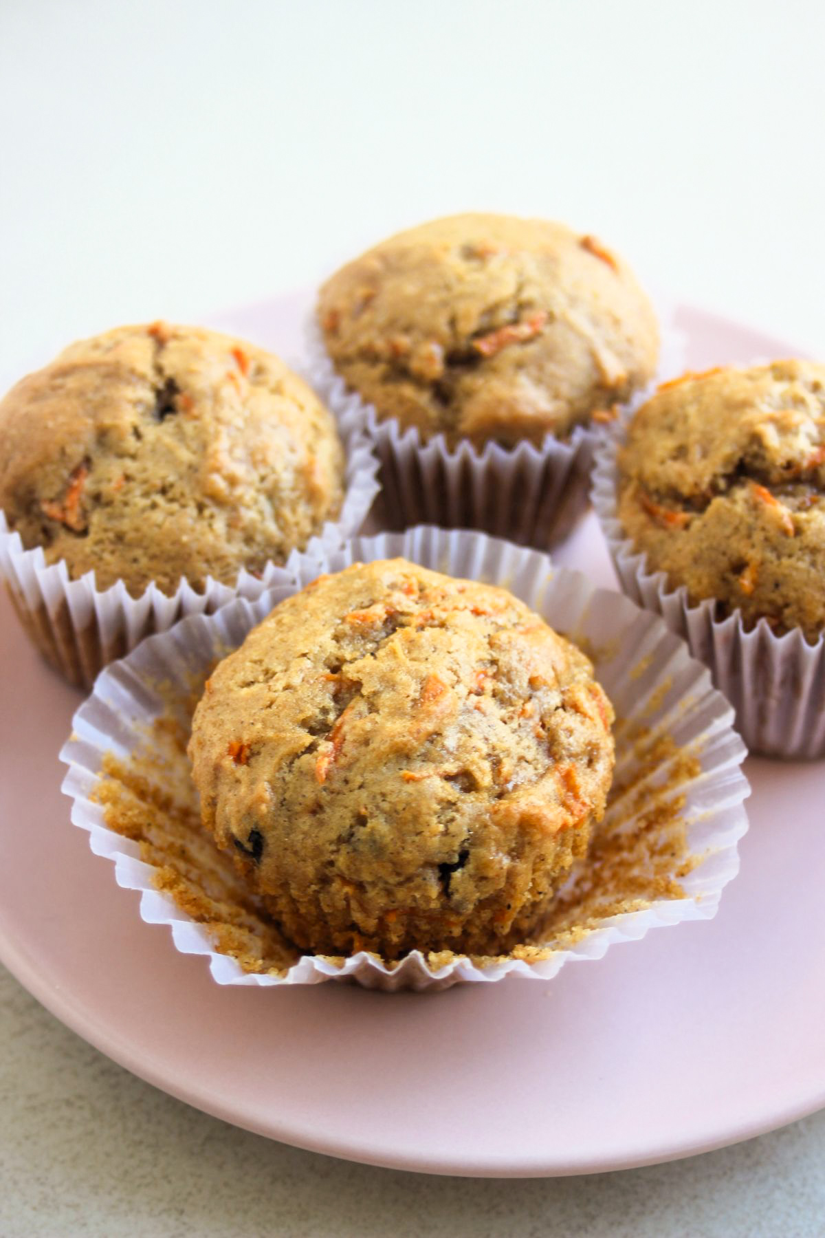 Carrot cake muffins in liners on a pink plate. The one in front, has the liner opened.