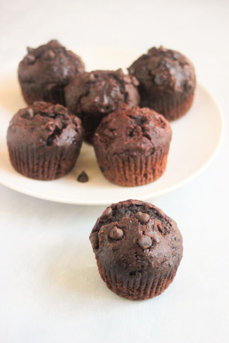 One chocolate beet muffin on a white surface. More muffins behind on a plate.