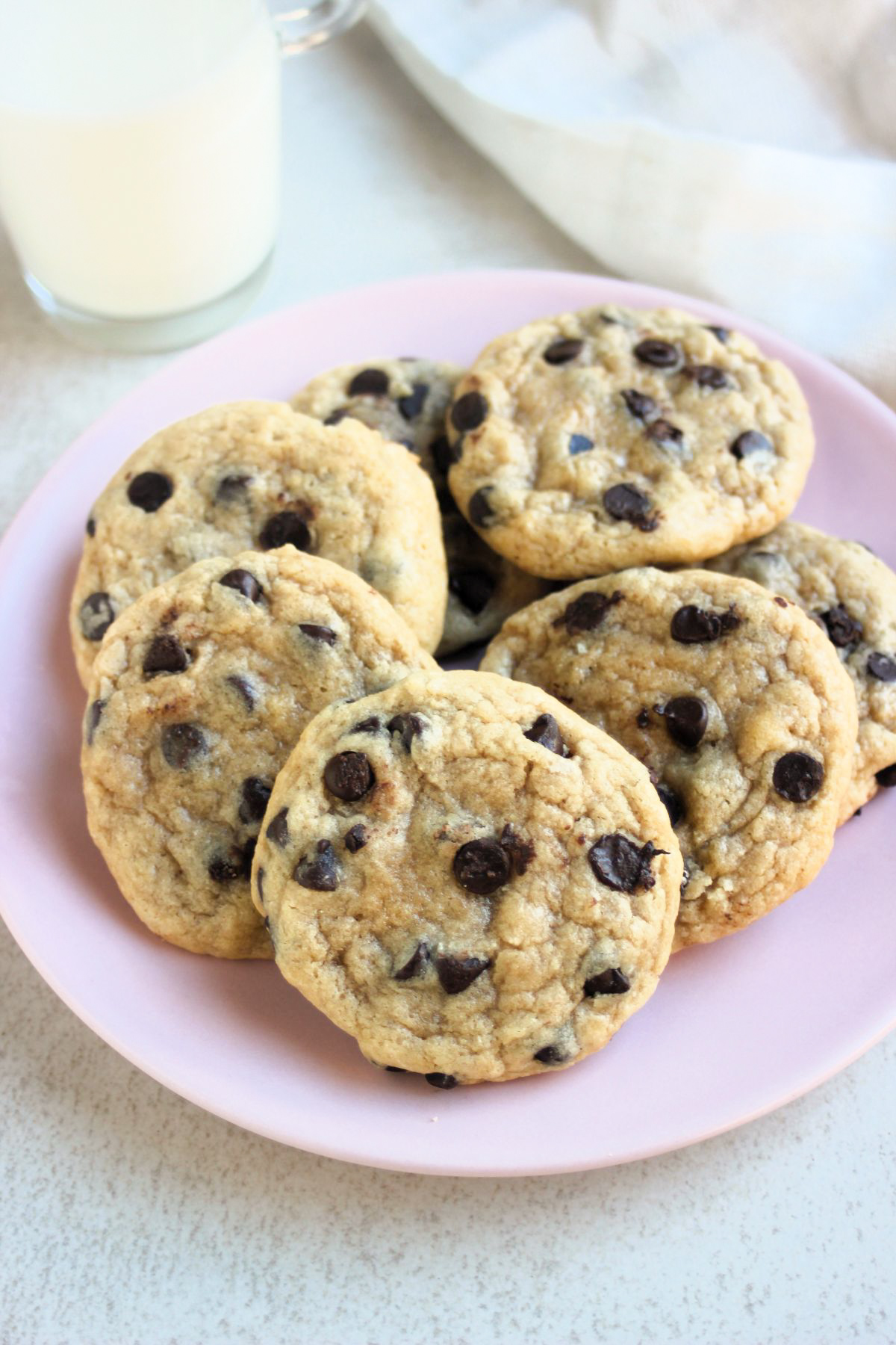 Chocolate chip cookies on a pink plate. White napkin and glass with milk behind.