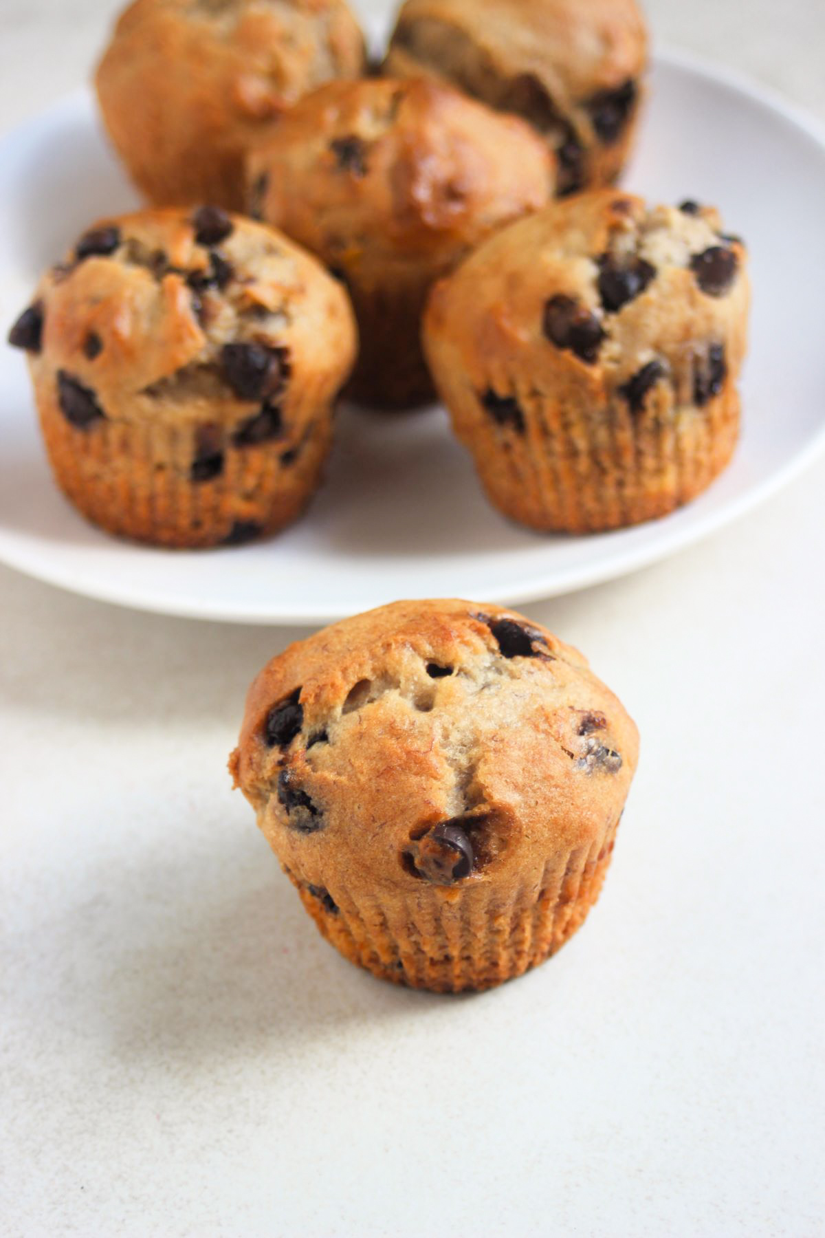 Banana chocolate chip muffin on a white surface. Behind, more muffins on a white plate.