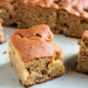 Portion of apple cake on a white surface. Whole cake behind.
