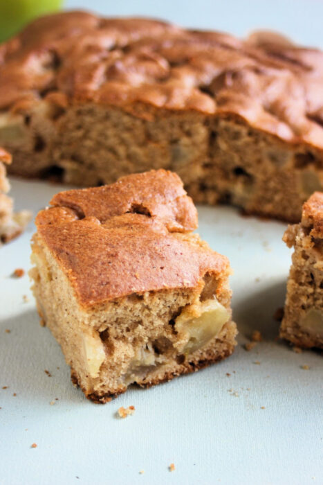 Portion of apple cake on a white surface. Whole cake behind.