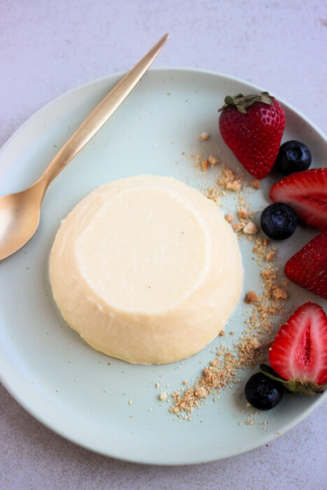 Top view of lemon panna cotta on an aqua green plate. A golden spoon, crumbs, fresh strawberries, and blueberries beside.