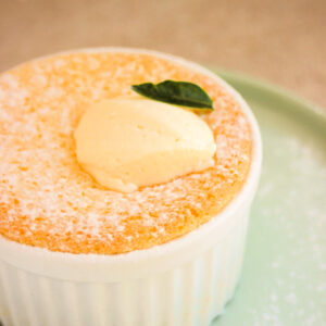 White ramekin with lemon pudding cake and whipped cream on an aqua green plate.