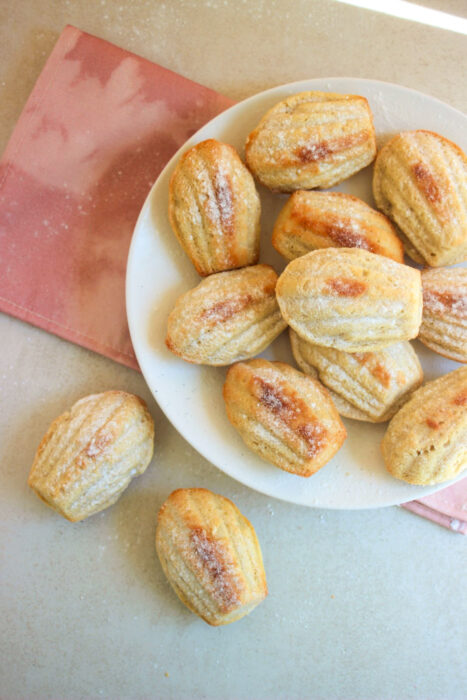 Many madeleines in a white plate, and two madeleines on the side. Pink napkin under the plate.