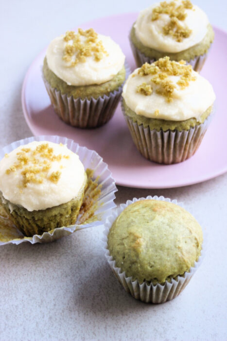 A matcha cupcake without frosting and a matcha cupcake with the liner open. Behind, more cupcakes on a pink plate.