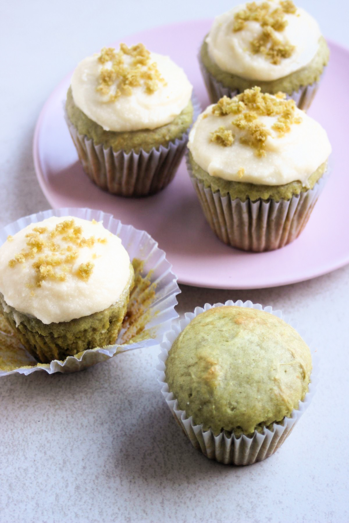 A matcha cupcake without frosting and a matcha cupcake with the liner open. Behind, more cupcakes on a pink plate.