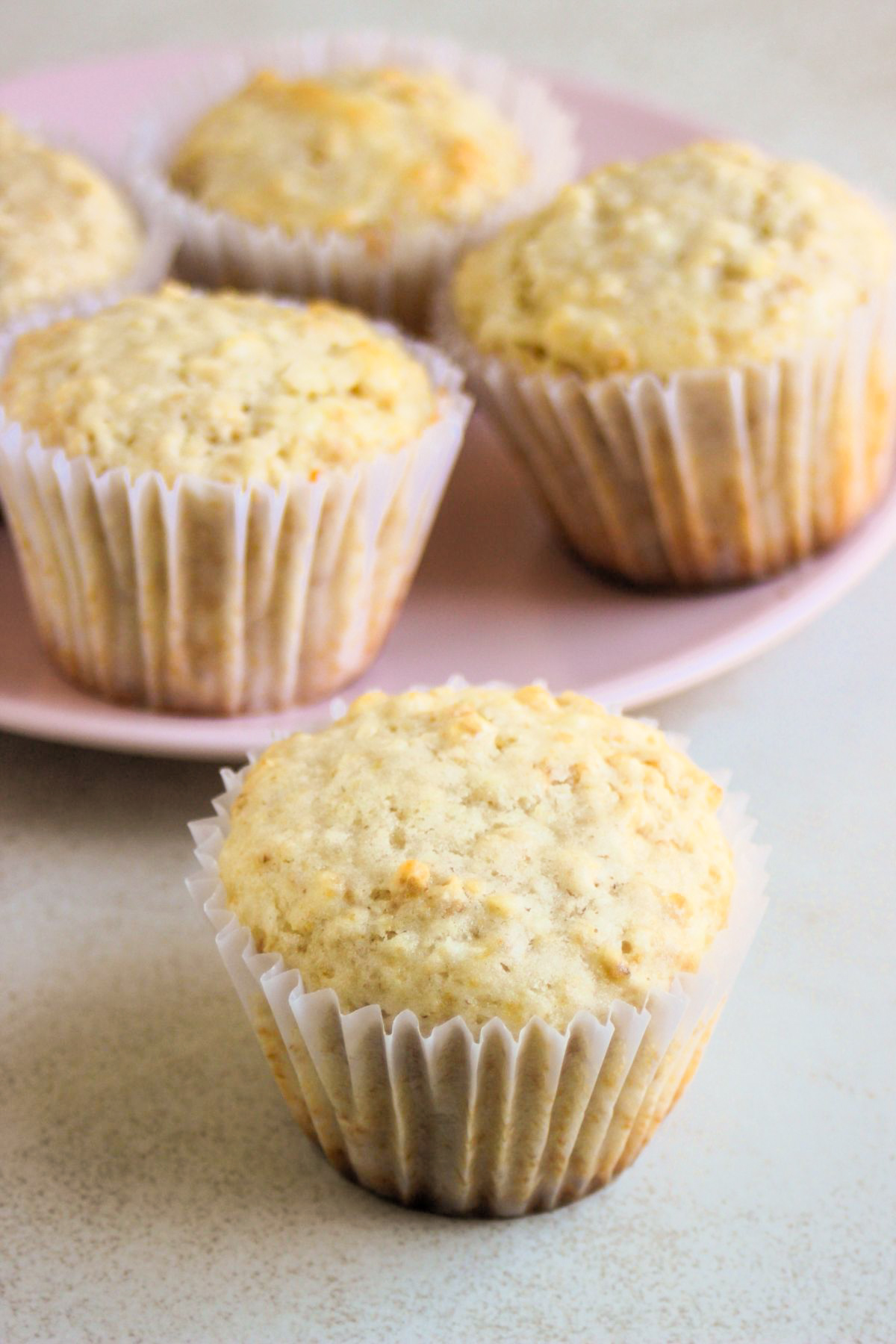 Oatmeal muffin with a liner on a white surface, more muffins behind on a pink plate.