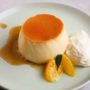 Orange flan with caramel, whipped cream, and fresh orange slices, on an aqua-green plate.