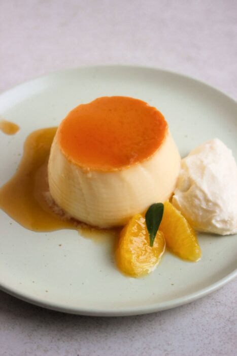 Orange flan with caramel, whipped cream, and fresh orange slices, on an aqua-green plate.