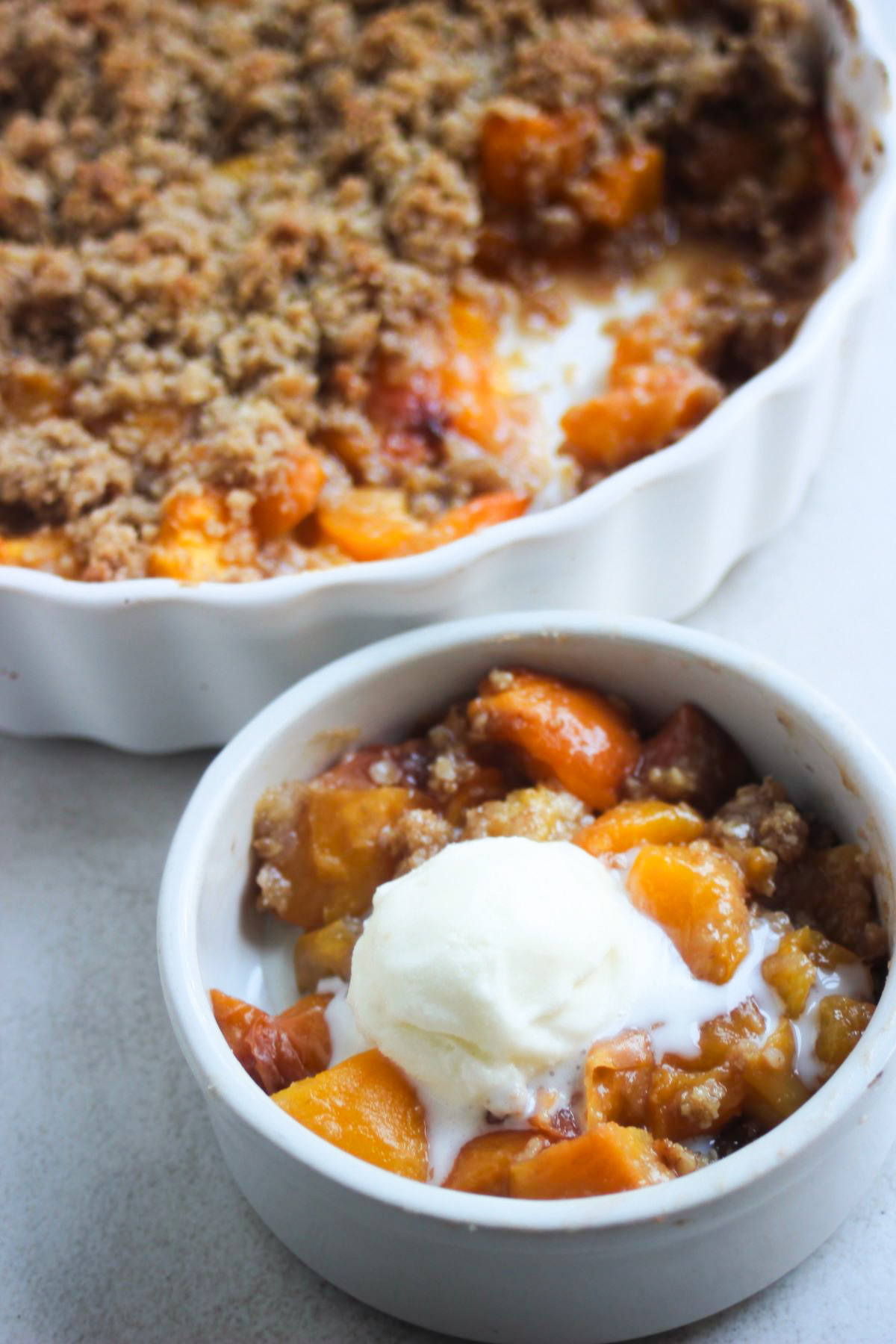 Serving of gluten-free peach crisp topped with vanilla ice cream. Baking dish with peach crisp on the background.