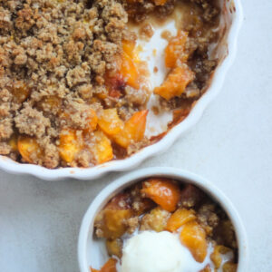 Small white ceramic bowl with peach crisp and a scoop of ice cream. White round baking dish with peach crisp behind.