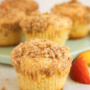 Peach streusel muffins on a white surface. Fresh peach slices on the sides, and more muffins behind on a plate.