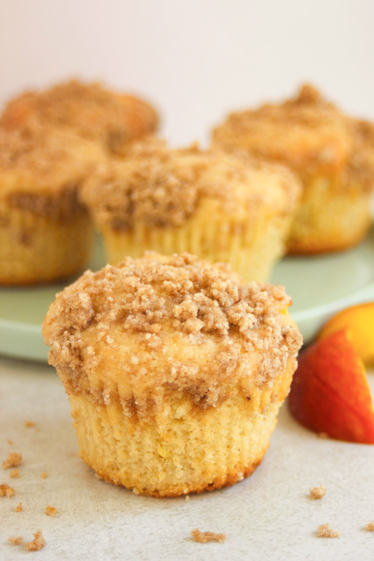 Peach streusel muffins on a white surface. Fresh peach slices on the sides, and more muffins behind on a plate.
