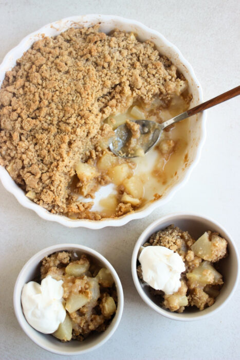 Two small bowls with pear crumble and whipped cream, and a round baking dish with pear crumble and a spoon seen from above.