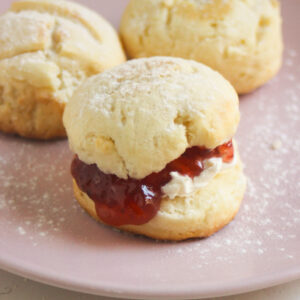 Scon filled with cream cheese and jam on a pink plate. More scones behind.