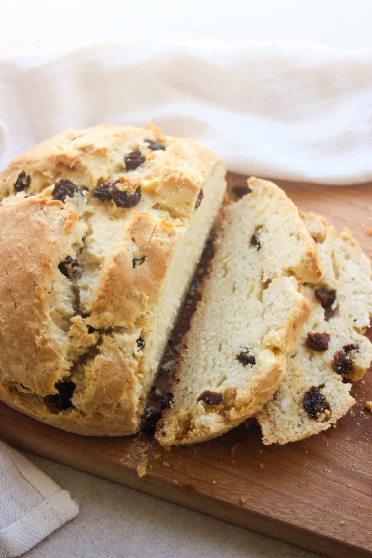 Soda bread with raisins and two slices of it on a wooden board. White kitchen towel behind.